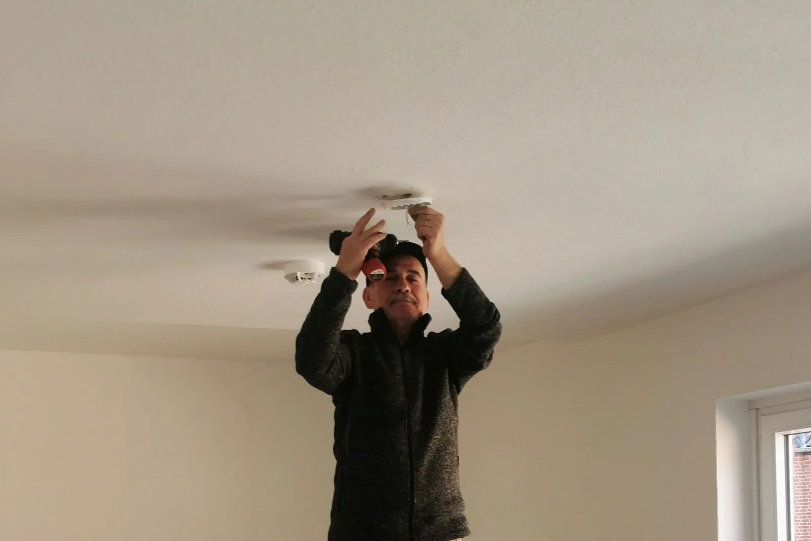 Man op een trapje werkt aan een montageplaat aan het plafond, met een rookmelder ernaast