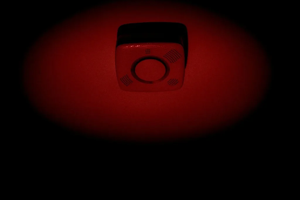 Rookmelder aan het plafond van een donkere kamer, in rood schijnsel; een zwakke batterij laat een melder vaak 's nachts piepen